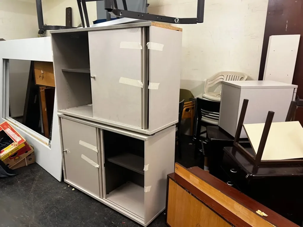 Zwei mit Klebeband gesicherte graue Büro-Sideboards, daneben Stapelstühle und Tischgestelle im Lager